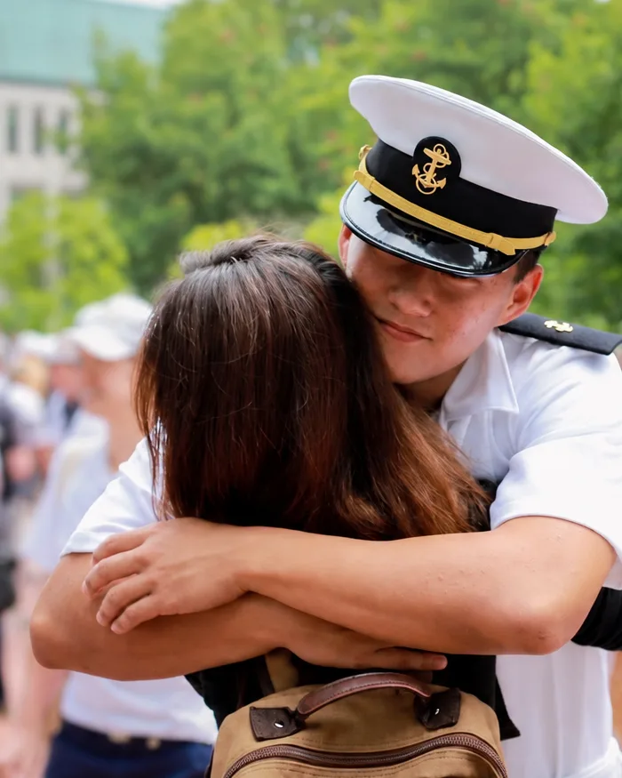 A Midshipman in summer whites hugs a parent, with other Mids and families gathered behind them.